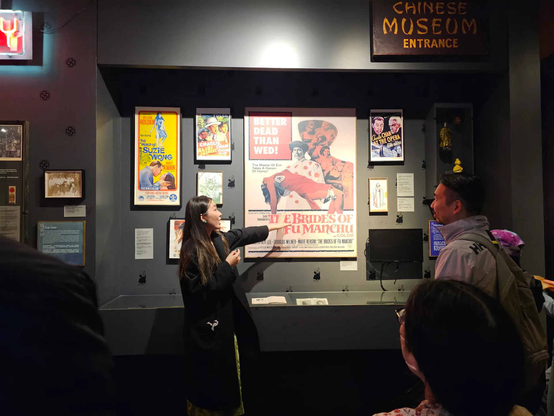 Gallery interior with exhibition wall displaying vintage Chinese American posters and signage, visitors engaged with a docent guide.