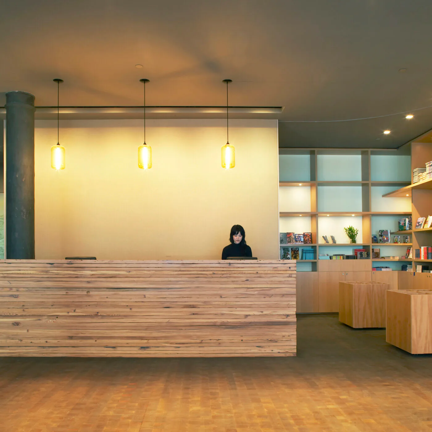 Modern museum reception area with light-wood counter, warm pendant lighting, built-in shelving, and staff member at desk.