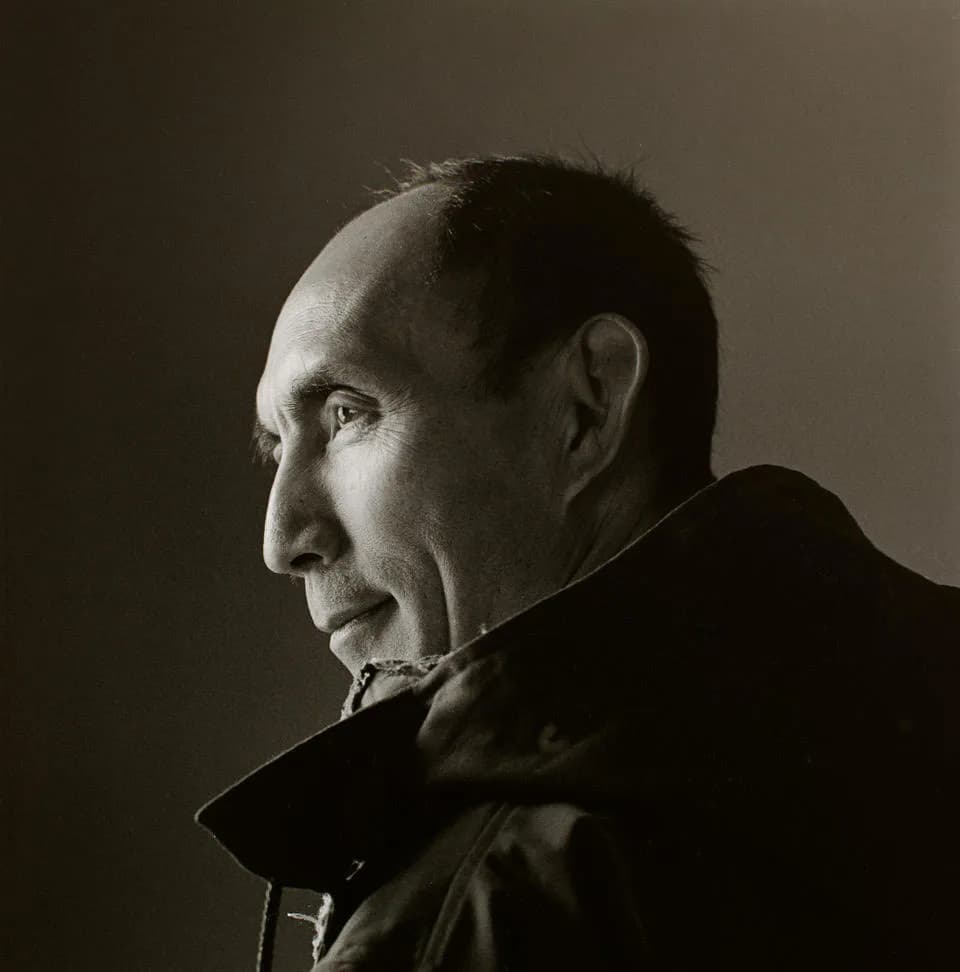 Black and white profile portrait of an older man in dark clothing against neutral background.
