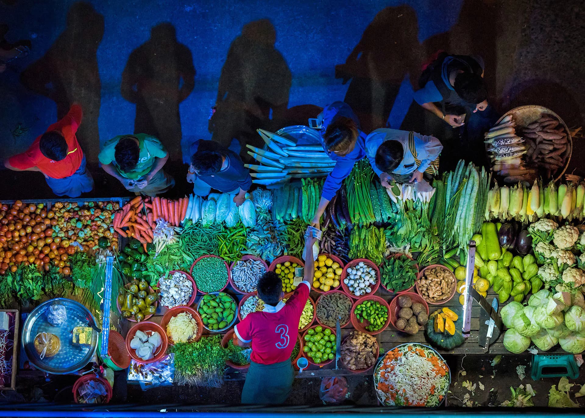 Overhead photograph of a vibrant produce market with neon-lit signage, vendors, and colorful fruits and vegetables arranged in circular patterns.