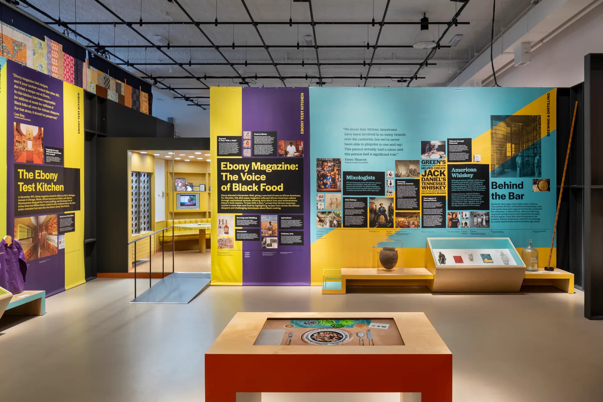 Interior of Museum of Food and Drink gallery with yellow, purple, and turquoise walls, educational panels, interactive displays, and a red exhibition platform.