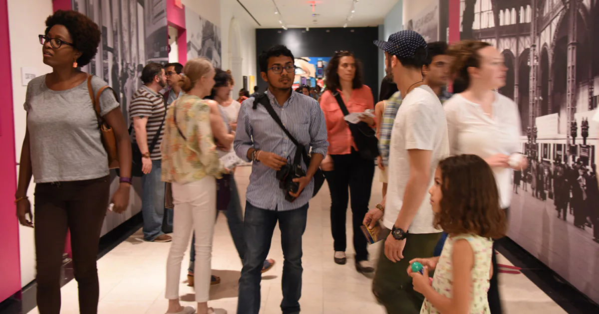 Museum gallery interior with visitors walking through a corridor lined with large architectural photographs on pink and dark walls.