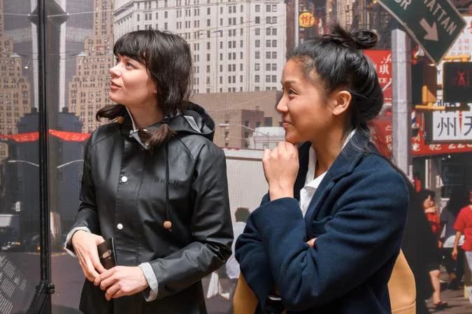Two visitors viewing a museum exhibition panel featuring Manhattan cityscape photography and text, with city skyline imagery visible in background.