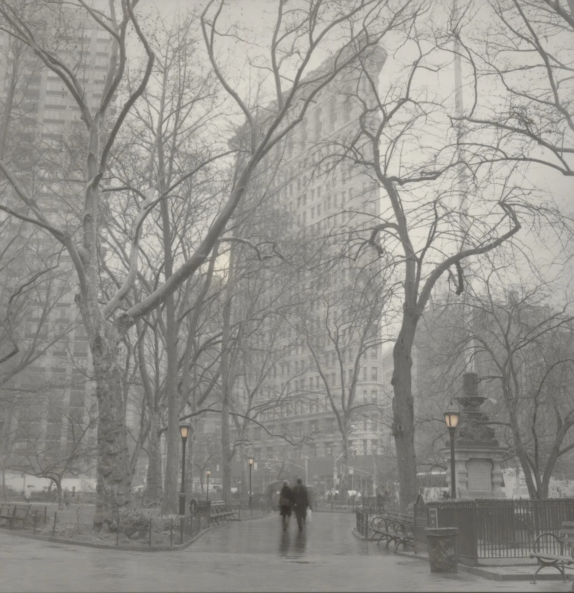 Black and white photograph of a tree-lined urban plaza with bare branches, classical architecture in background, two figures walking in misty atmosphere.