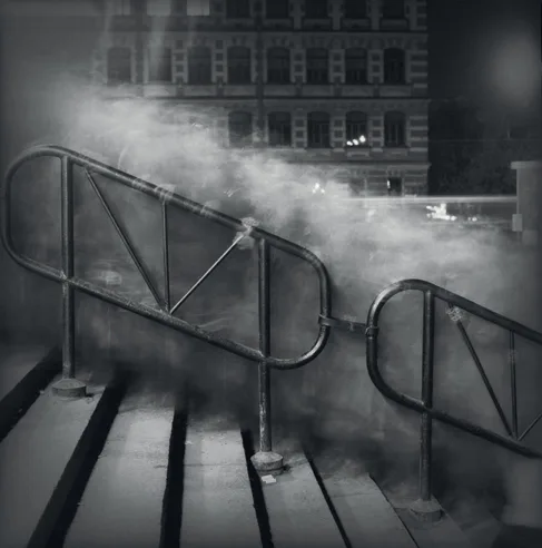 Black and white photograph of metal handrails and steps on a rooftop with steam or smoke rising, urban building facade behind.