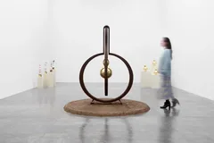 Minimalist sculpture featuring a large black circular metal ring mounted on a natural fiber base, displayed in a white gallery space with candles nearby.