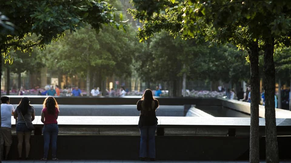 Visitors viewing the memorial reflecting pool framed by mature trees with dappled sunlight.