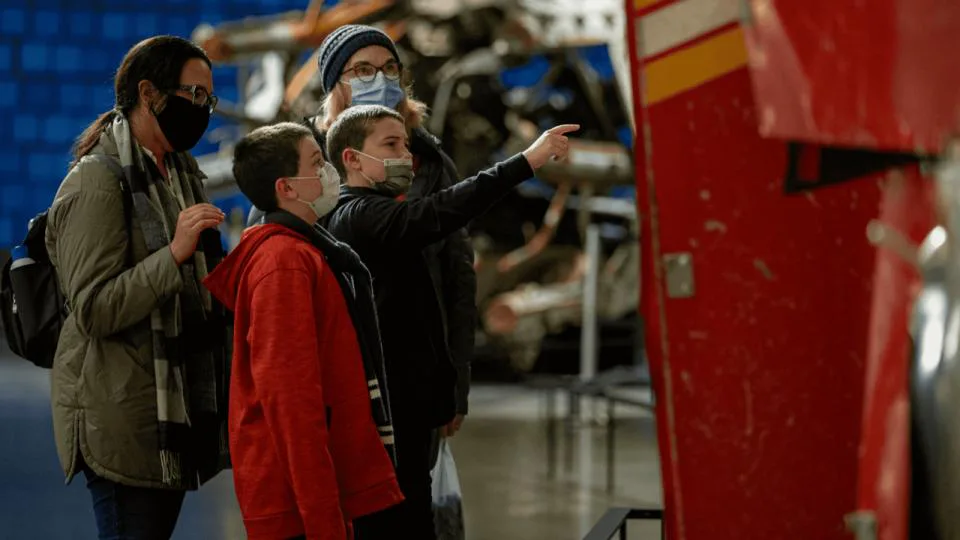 Museum visitors, including children, looking at exhibits with machinery visible in background.