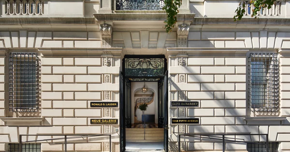 Storefront exterior of Neue Galerie New York with cream-colored stone facade, black entrance door with ornate ironwork, and symmetrical window grilles flanking the entry.
