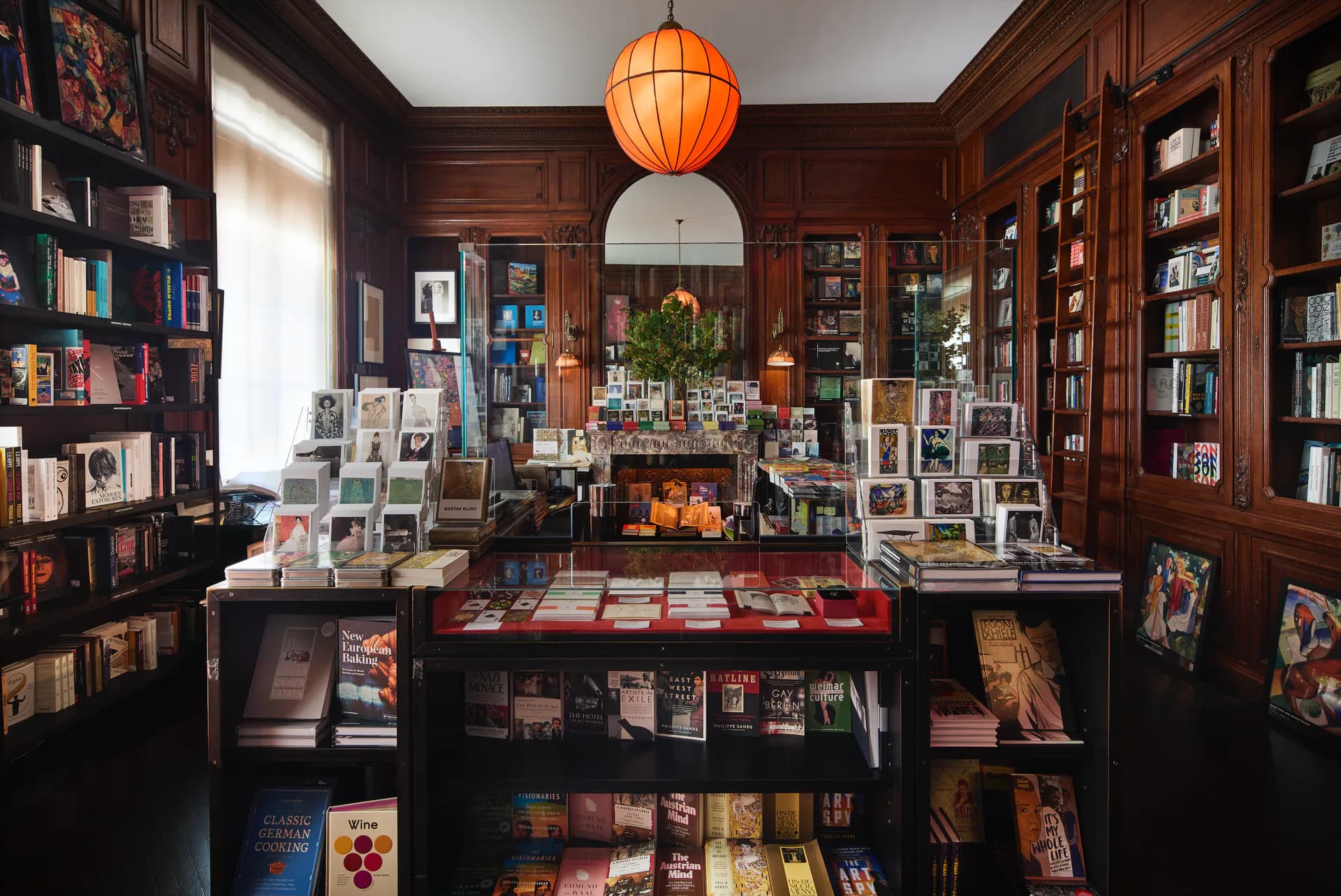 Interior of Neue Galerie bookstore with dark wood paneling, floor-to-ceiling shelves, central display table with books and artworks, and an orange paper lantern pendant light.