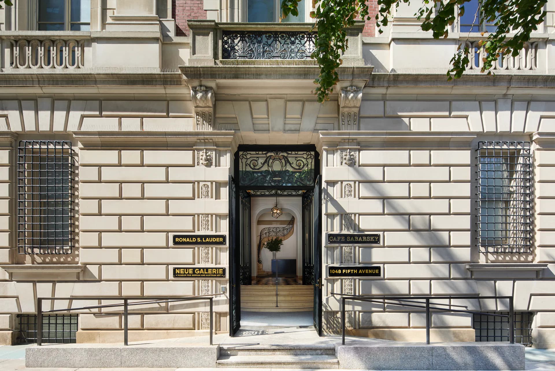 Neue Galerie storefront with cream stone facade, centered black ornate entrance door, decorative window grilles, and shop signage on the building exterior.
