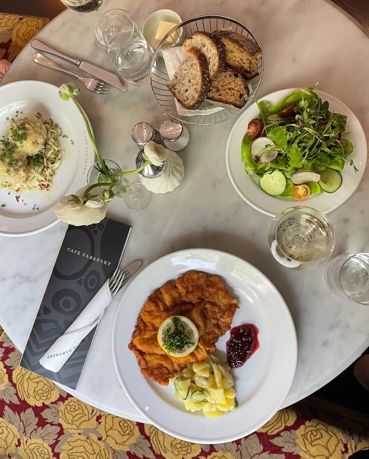 Overhead view of a restaurant table with plated dishes including schnitzel and salad, bread basket, water glasses, and a café menu on marble surface.