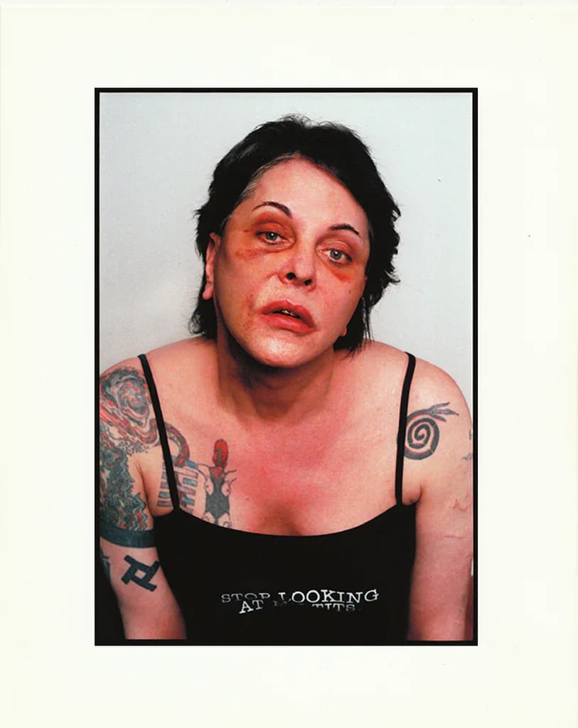 Portrait photograph of a person with dark hair and visible tattoos on their arms, against a white background.