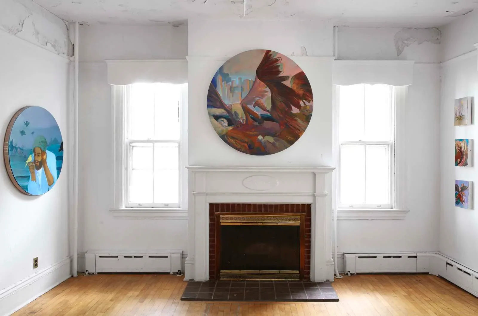 Gallery room with fireplace, two windows with natural light, wooden floor, circular paintings mounted on walls above fireplace and to the side, featuring figural compositions.