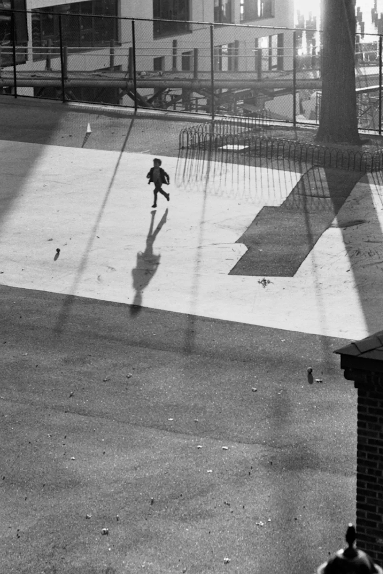 Black and white photograph of child running across sunlit concrete plaza with strong geometric shadows from railing and tree.