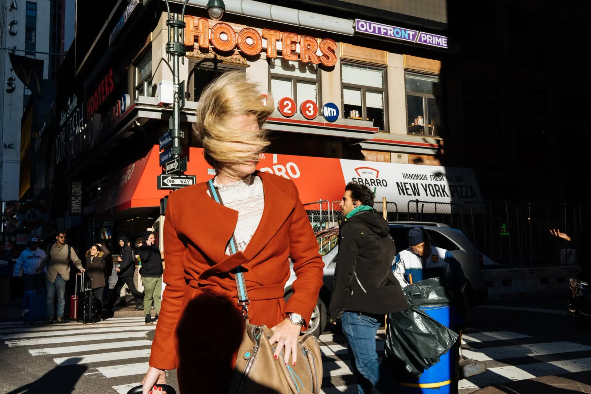 Color photograph of blonde woman in bright red jacket walking through Times Square with neon signage and crowds in background.