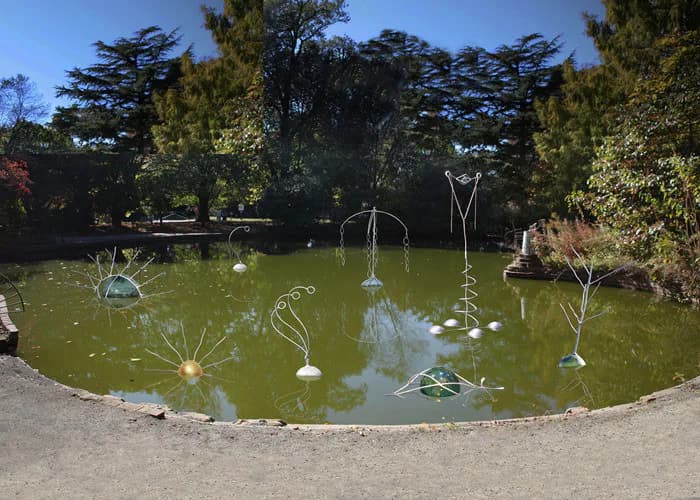 Installation artwork in a circular pond featuring suspended wire and metal sculptures with hanging spheres and organic forms against lush greenery.
