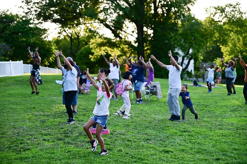 Children and adults engaged in recreational activities on a grassy lawn surrounded by mature trees at Snug Harbor.