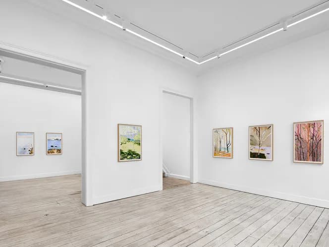 Minimalist gallery interior with light wood flooring, white walls, central pedestal, and small framed artworks displayed at varying heights.