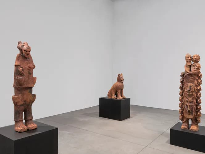 Gallery space with three wooden carved figurative sculptures mounted on black pedestals against white walls.