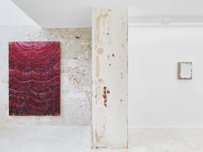 Raw gallery space with textured walls, distressed wooden panel divider, magenta-red patterned textile artwork, and small framed piece on wall.