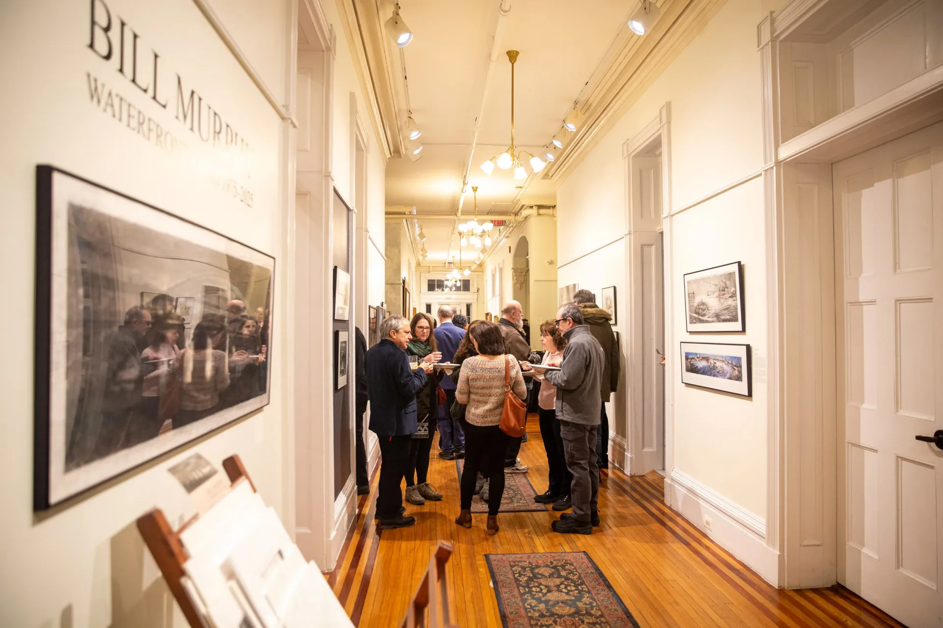 Interior of Noble Maritime Collection gallery: narrow hallway with framed photographs on white walls, visitors viewing exhibits, warm lighting and wooden floors.
