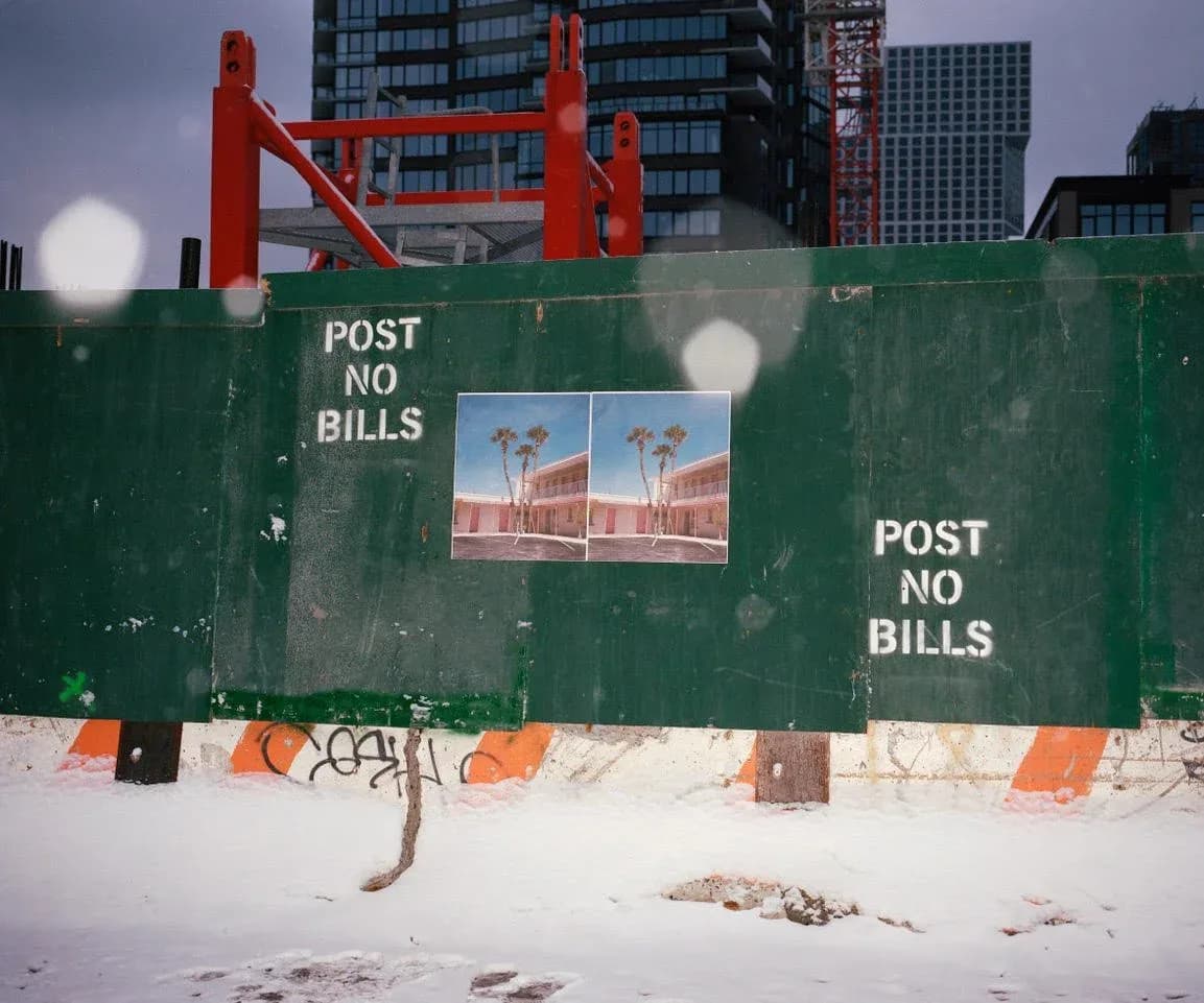 Photograph of a snowy urban construction site with a green barrier wall bearing 'POST NO BILLS' text, tall buildings visible behind.