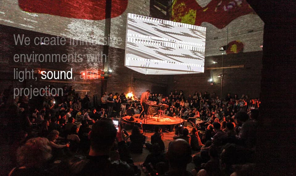 A seated audience surrounds a circular stage with musicians as an abstract black-and-white pattern is projected on a large screen above, at Nuit Blanche New York.