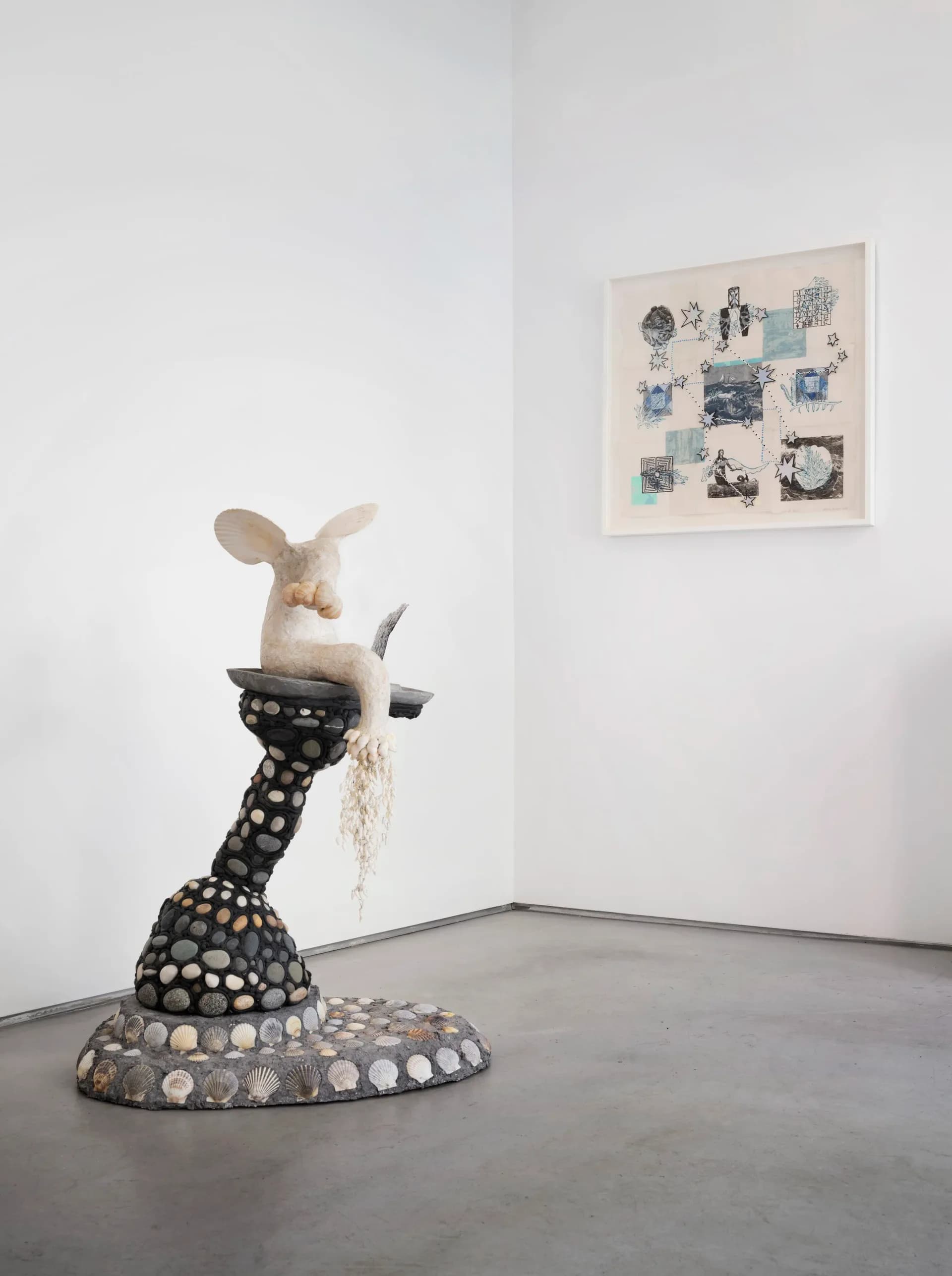 Gallery corner with pale ceramic sculptural figure on decorative black pedestal base and framed mixed-media artwork on wall above.