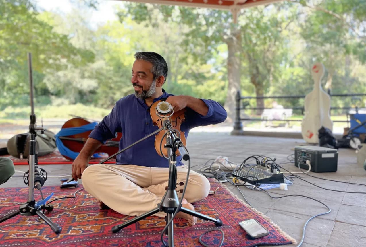 Man seated outdoors on a patterned rug holding a stringed instrument, wearing a blue shirt, with trees and park setting visible in background.