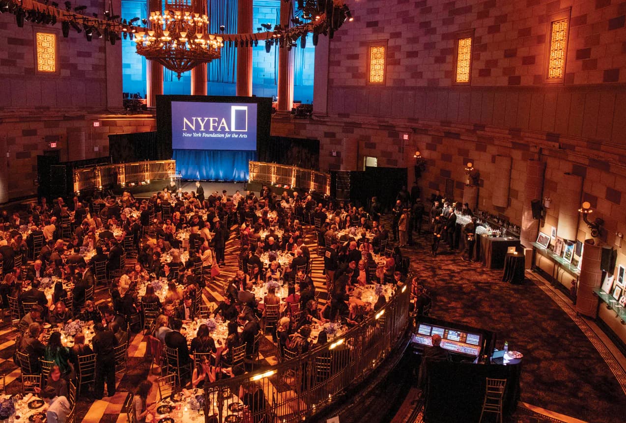 Interior of NY Foundation for the Arts venue during an event: crowded ballroom with red brick walls, chandeliers, illuminated NYFAL sign, and numerous attendees at tables.