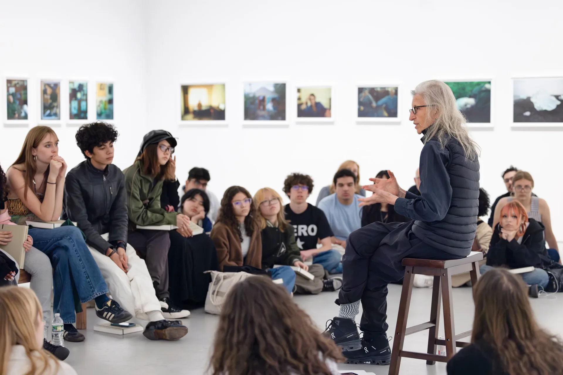 Gallery interior with seated audience facing a woman standing on a stool during what appears to be a presentation or artist talk, with framed photographs on white walls.