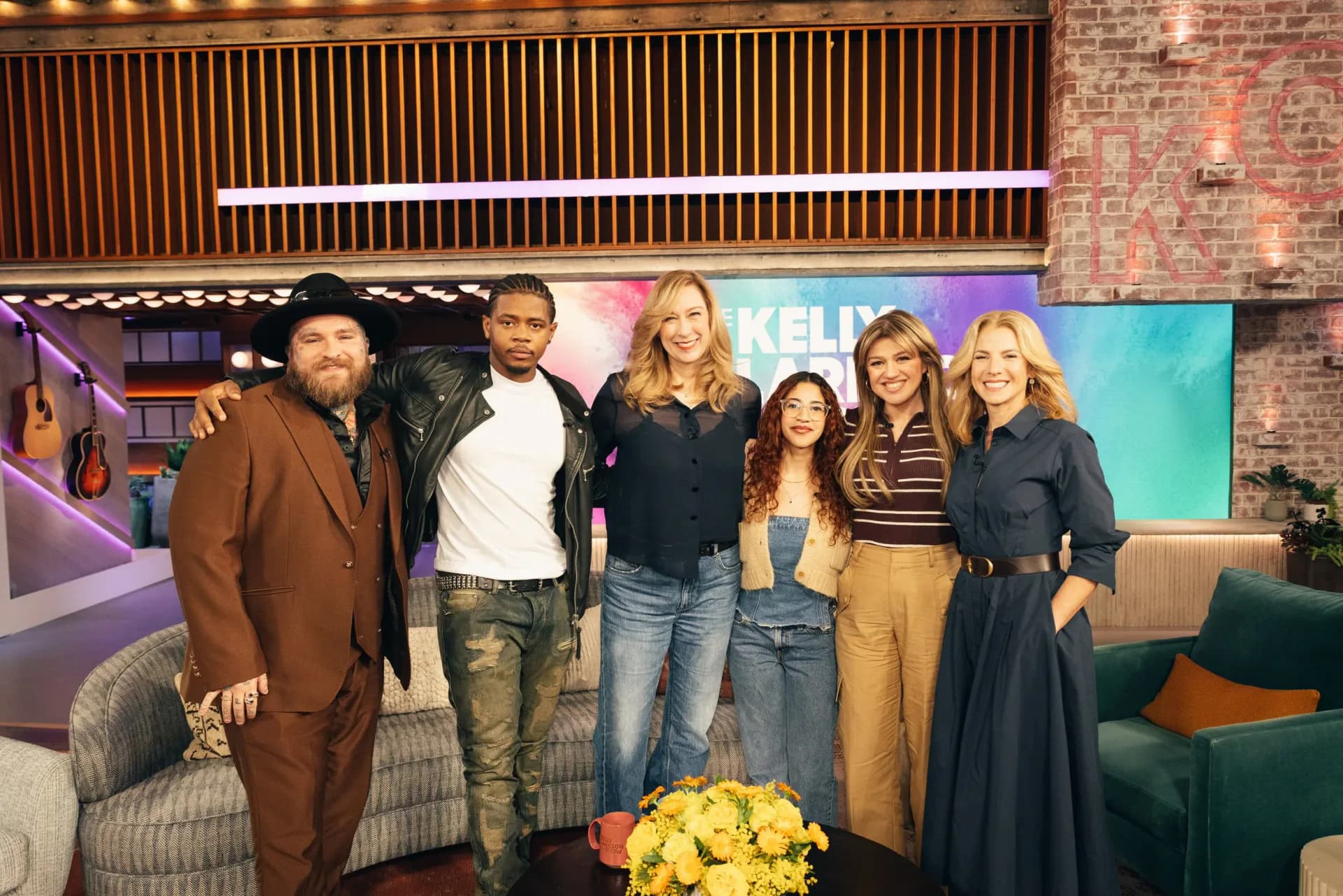 Group of six people posing together on a television studio set with colorful gradient background and yellow flowers in foreground.