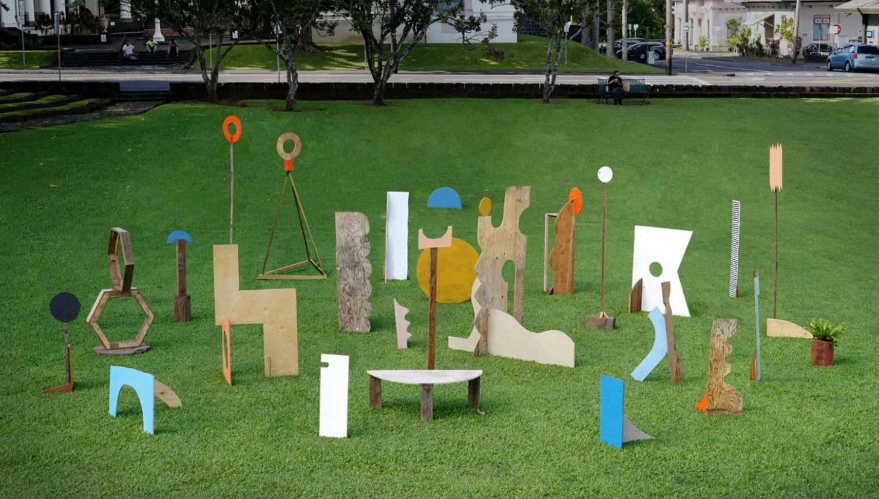 Large-scale outdoor installation on grass with mixed wooden, metal, and painted sculptural forms arranged in abstract composition.