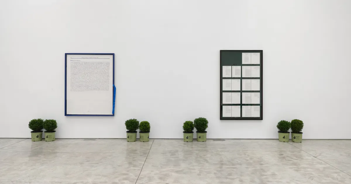 Gallery interior with white walls, concrete floor, and four potted green plants spaced across the floor, with a blue-framed white canvas on the left and a dark grid artwork on the right.