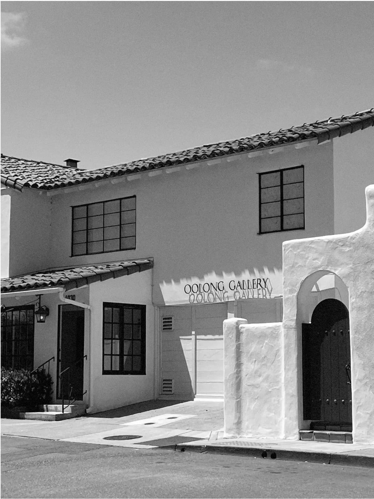 Black and white photograph of Oolong Gallery's exterior: white-walled building with dark windows, clay tile roof, and arched doorway entrance.