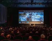 Theater auditorium with rows of seated audience members facing a large projection screen displaying an aerial urban photograph.