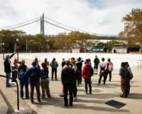 Outdoor waterfront plaza with a group of visitors standing near a green suspension bridge visible in the background under cloudy sky.