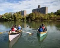 Waterfront scene with multiple kayakers in colorful boats on the river, downtown skyline and tree-lined banks visible in daylight.