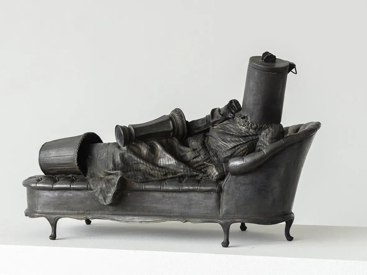 Dark cast-iron sculptural piece depicting a reclining figure on ornate legs, with various objects arranged on and around the form.
