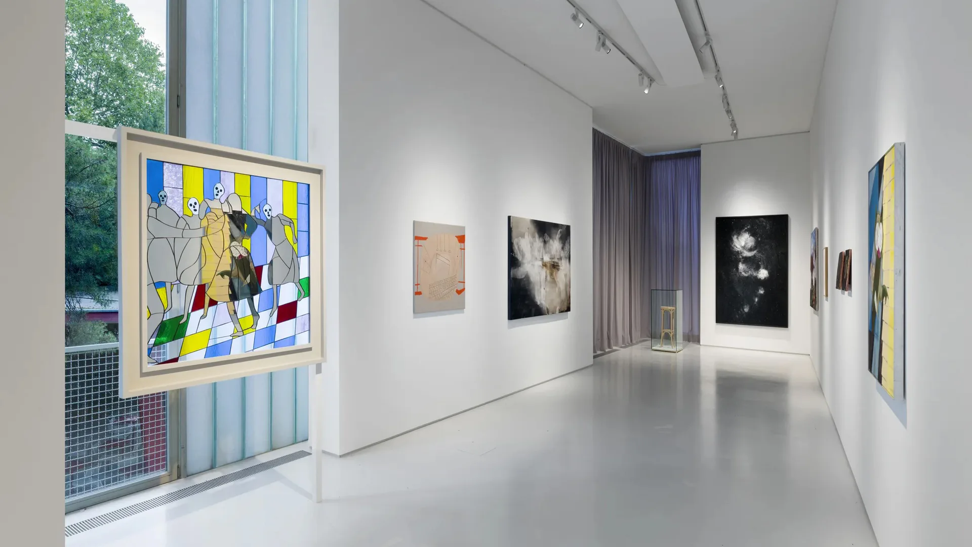 Bright gallery interior with polished white floor and white walls displaying framed artworks; colorful geometric painting visible on left, darker portraits and abstract works on right walls.