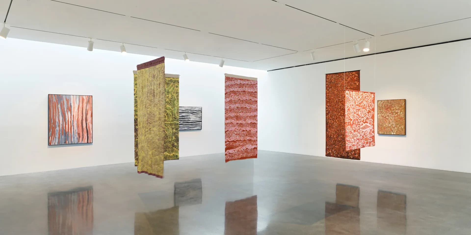 Gallery interior with freestanding colored panels in earth tones (coral, mustard, rust) and smaller artworks on white walls, polished concrete floor.