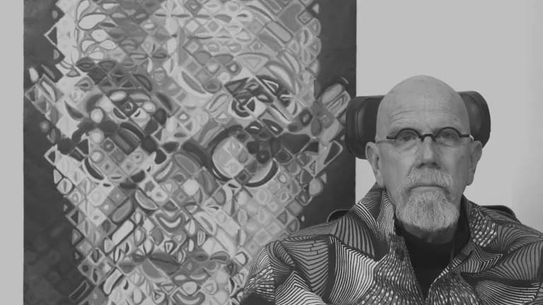 Black-and-white photograph of an older man with glasses and facial hair wearing a striped shirt, positioned next to an abstract textured artwork.