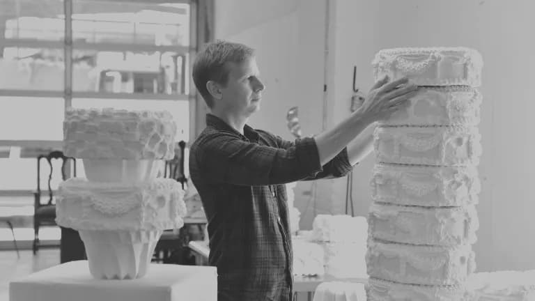 Black-and-white photograph of a person from side profile reaching toward a tall stacked sculptural form made of white geometric components in a studio setting.