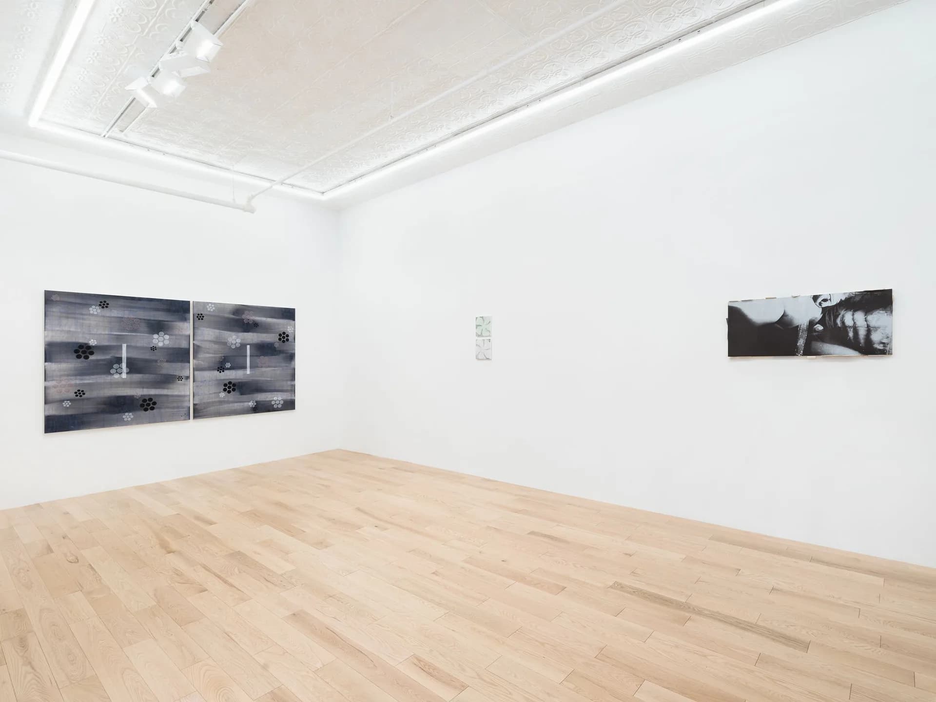 Gallery interior with light wood floors, black and white diptych on left wall, small black and white photograph on right wall, white ceiling with recessed lighting.