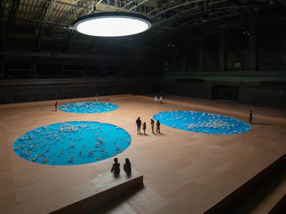 Large gallery interior with polished wood floor, three bright blue circular installations, overhead skylight, and visitors walking through the space.