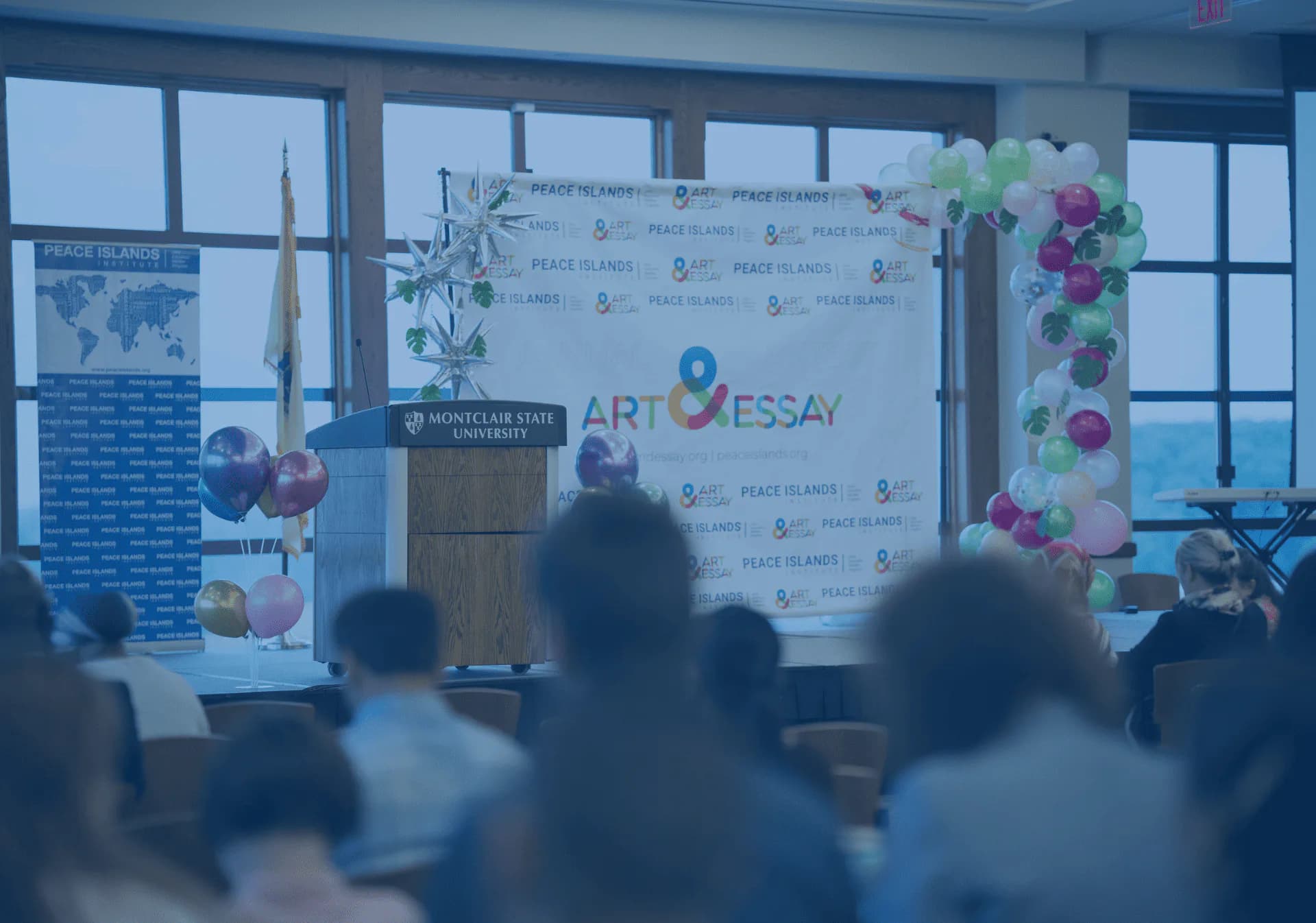 Conference room with attendees seated, podium with backdrop featuring Art Essay branding, large windows, and floral decorations.