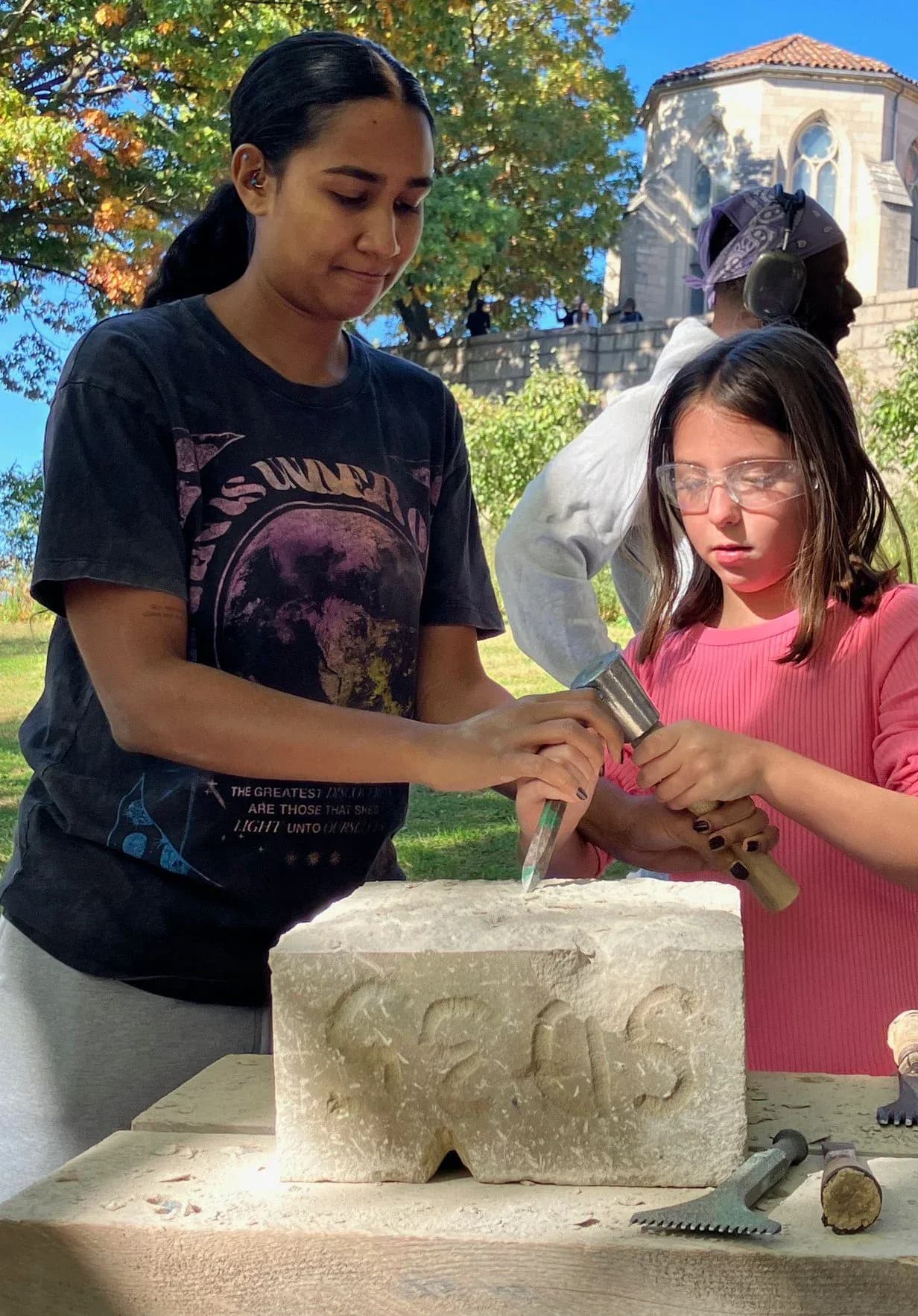 Two people, one guiding a young girl's hands, carving letters into a stone block outdoors near a historic building at Pellettieri Stone Carvers' Academy