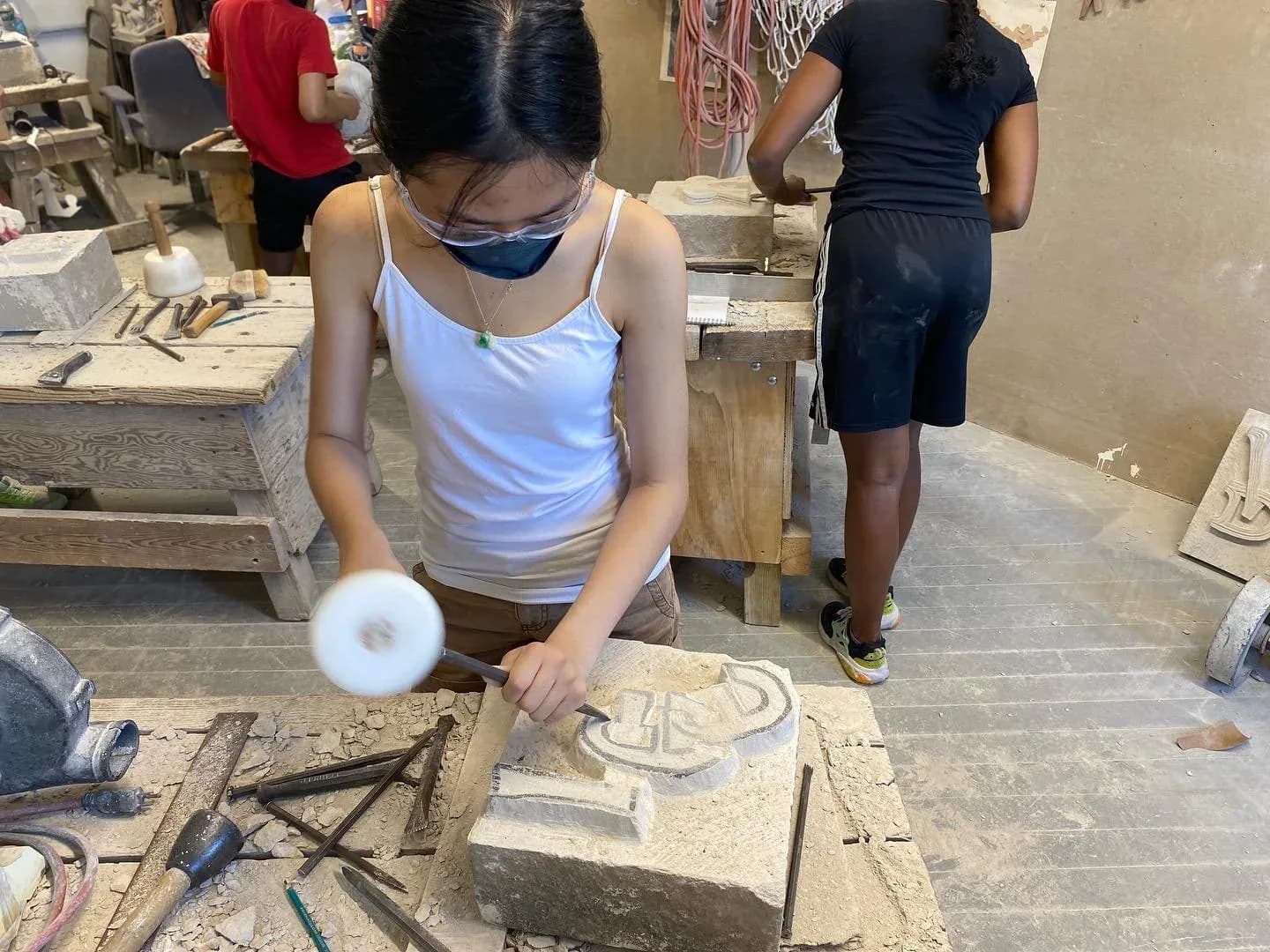 Young woman wearing a mask and safety glasses hammering a chisel into a carved stone relief in a workshop at Pellettieri Stone Carvers' Academy