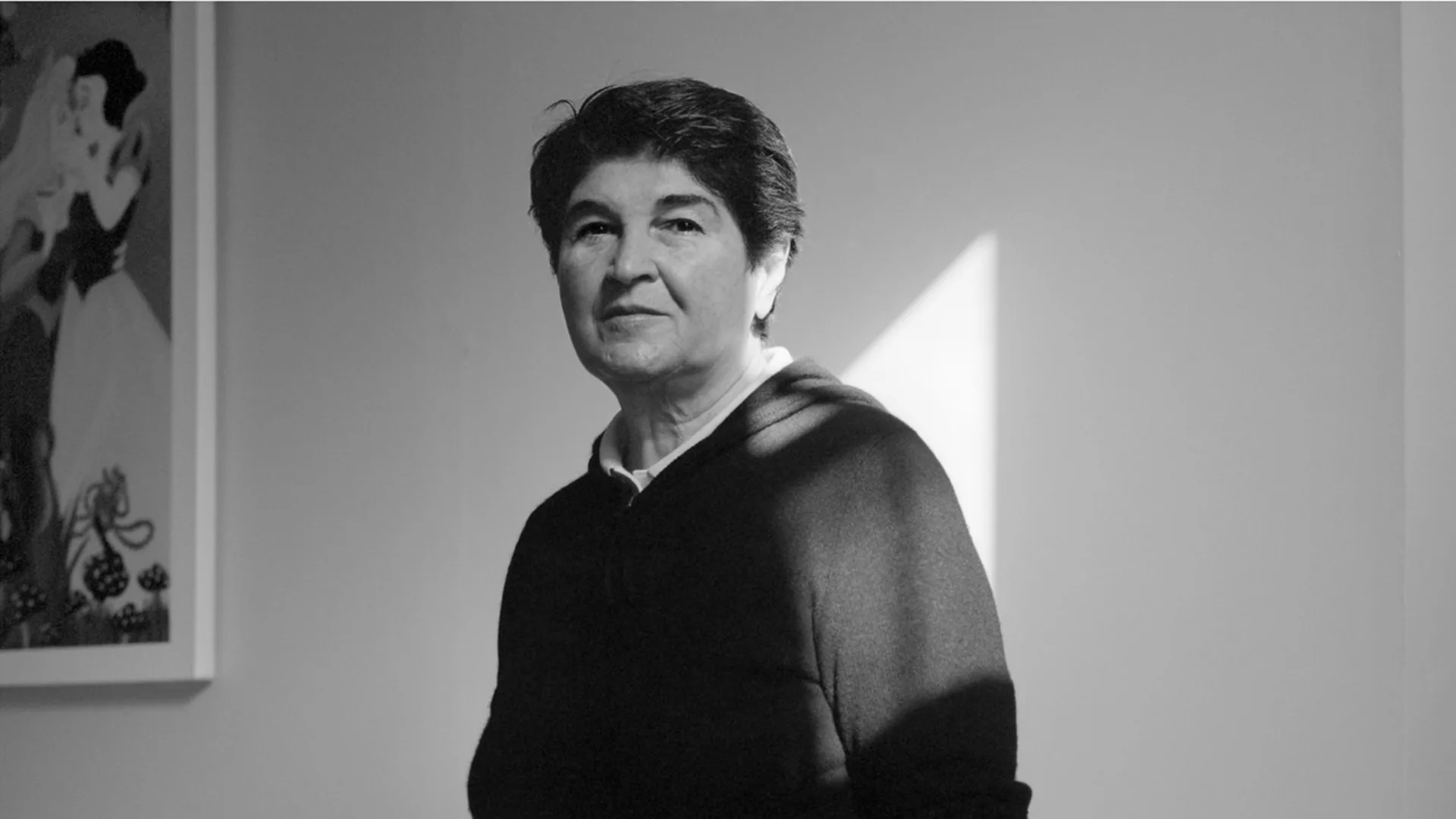 Black and white portrait photograph of a middle-aged person in dark clothing against white wall with framed artwork visible.